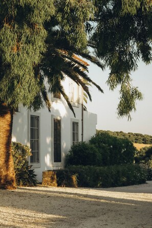A white building with barred windows, surrounded by greenery and a gravel path.