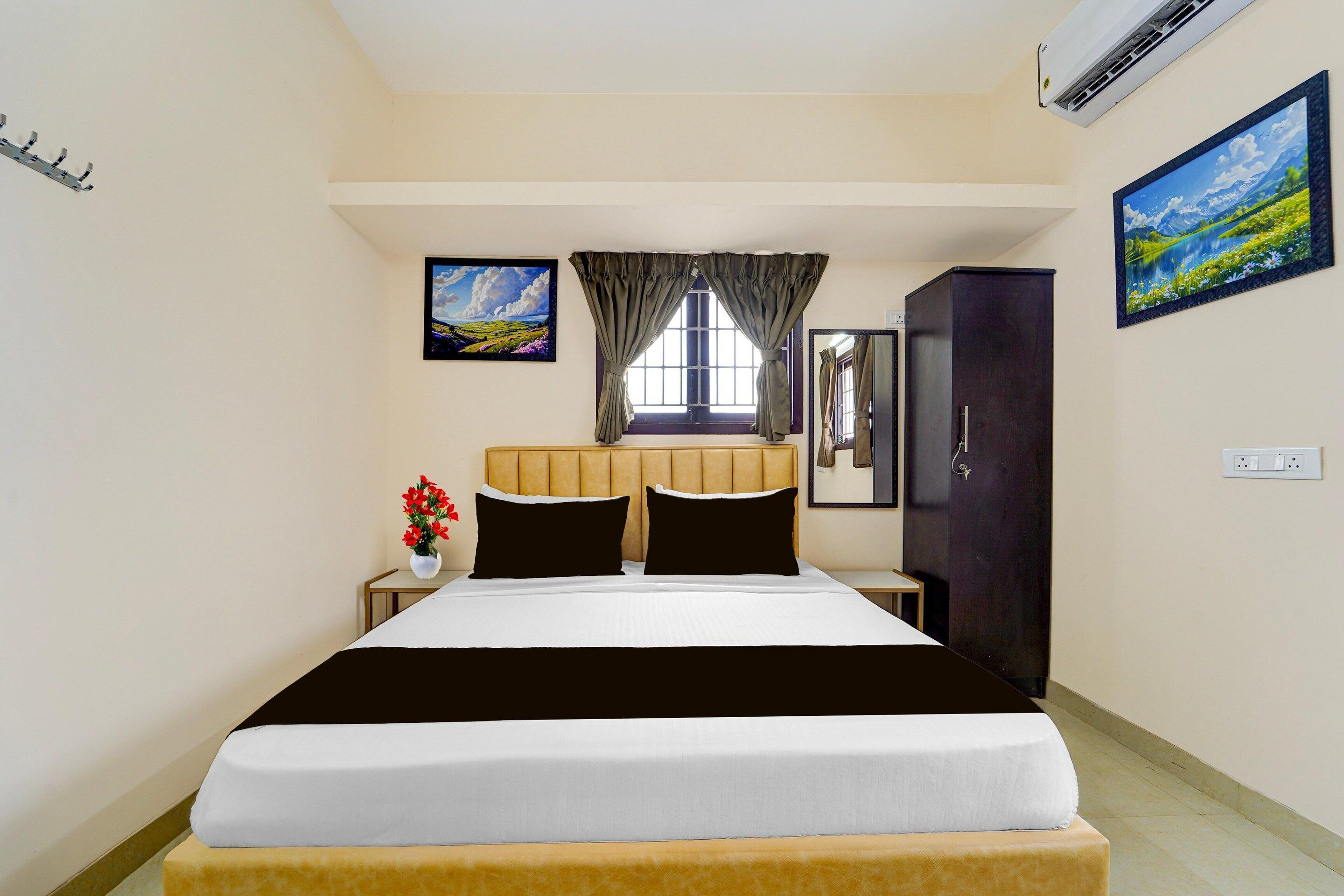 A hotel room with a bed, a window with curtains, a painting on the wall, a mirror, and an air conditioning unit.