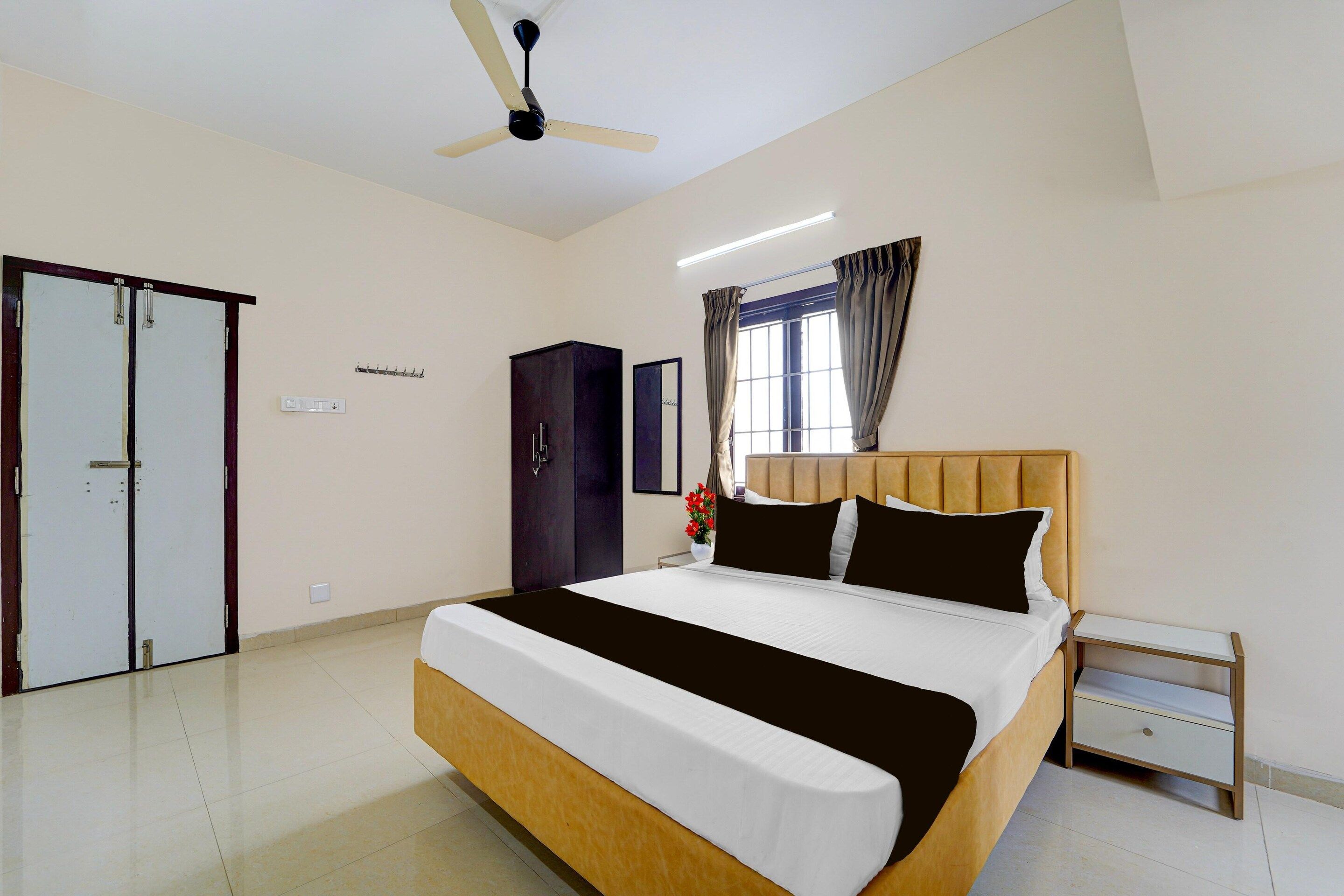 A hotel room with a bed, a nightstand, a wardrobe, a ceiling fan, and a window with curtains.