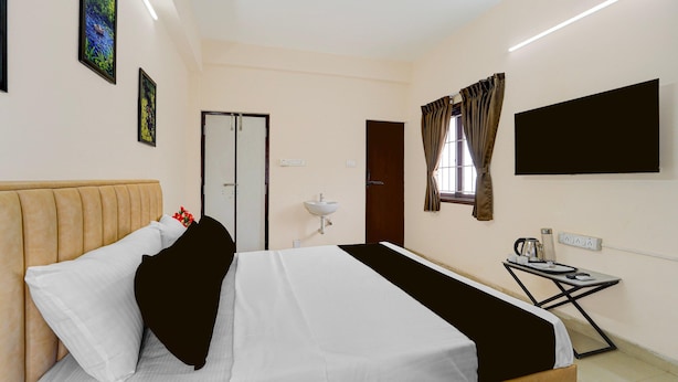 A hotel room with a bed, a bedside table with a kettle, a flat-screen TV, and a window with curtains.