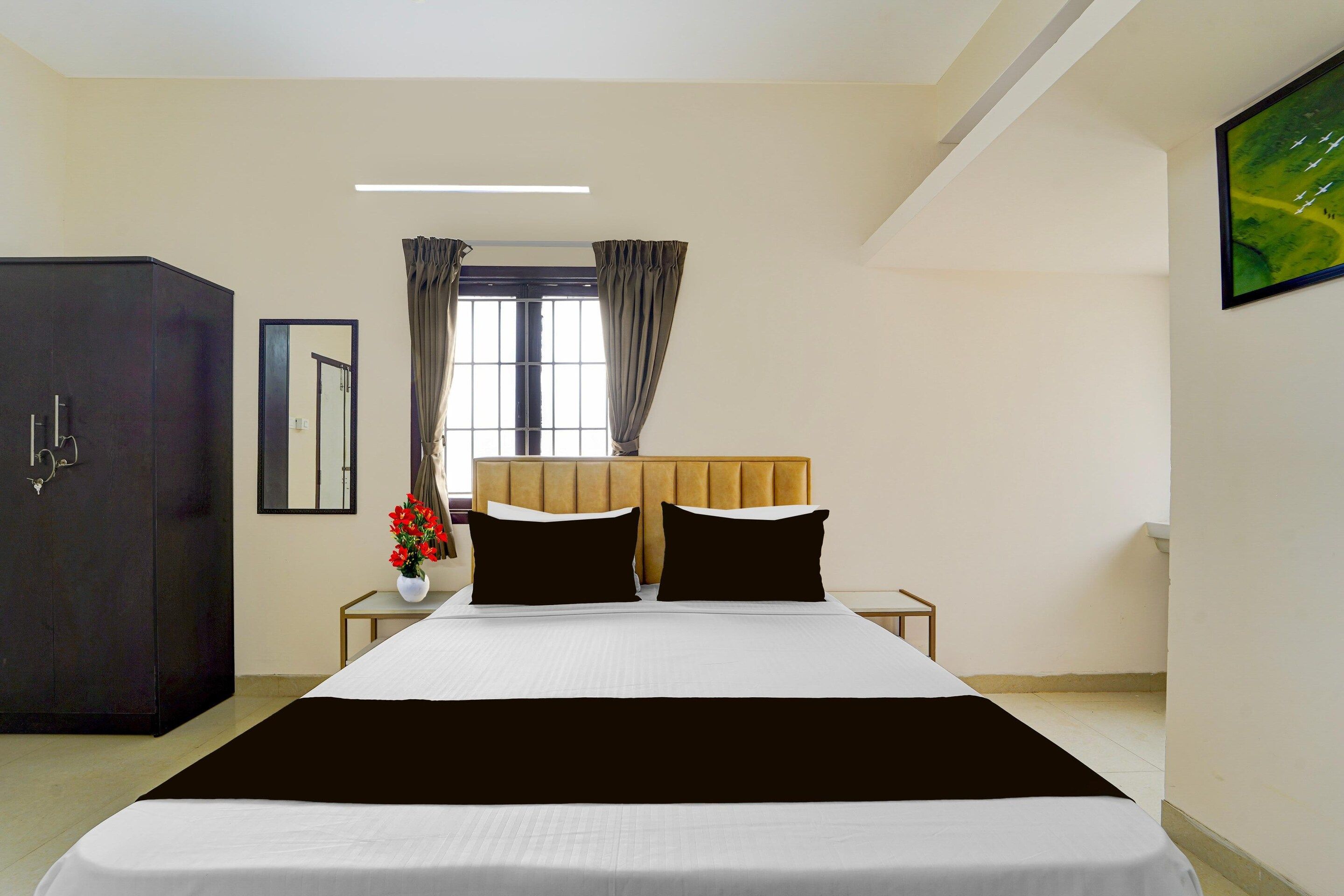 A hotel room with a bed, a wardrobe, a nightstand, a vase with flowers, and a window with curtains.
