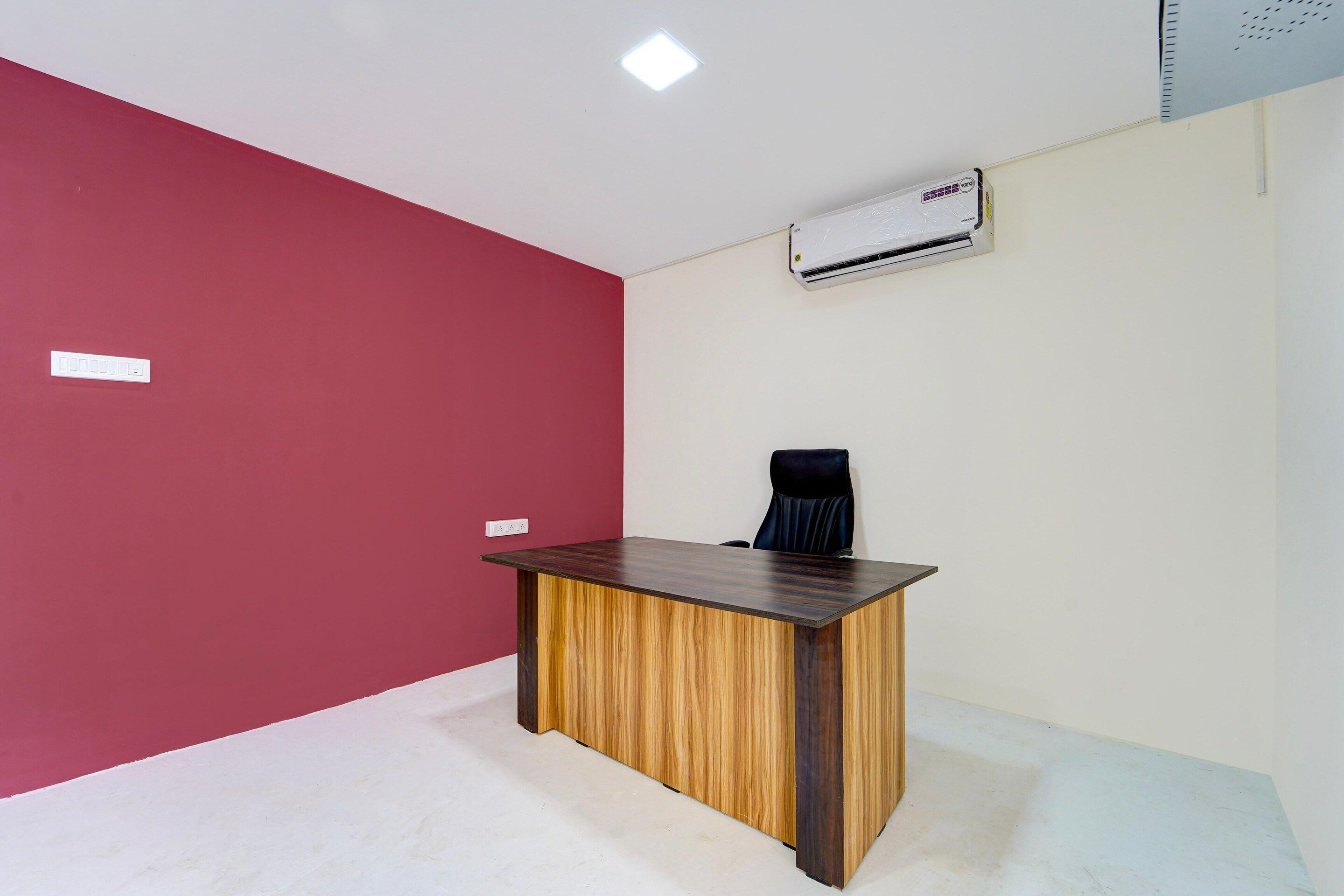 A room with a wooden desk, a black office chair, a white air conditioning unit on the wall, and a red accent wall.