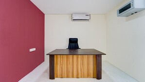 A room with a wooden desk, a black office chair, a white air conditioning unit, and a white wall.