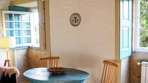 Une pièce avec une table ronde, deux chaises, un bol posé sur la table, une chaise en bois, un tapis, une applique murale et une fenêtre avec des volets bleus.