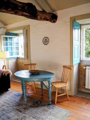 Ein Zimmer mit einem runden Tisch, zwei Stühlen, einer Schüssel auf dem Tisch, einem Holzstuhl, einem Teppich, einer Wandplatte und einem Fenster mit blauen Rollläden.