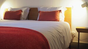 Un lit bien fait, avec une couverture rouge et des oreillers, une table de chevet avec une lampe et une prise électrique au mur.