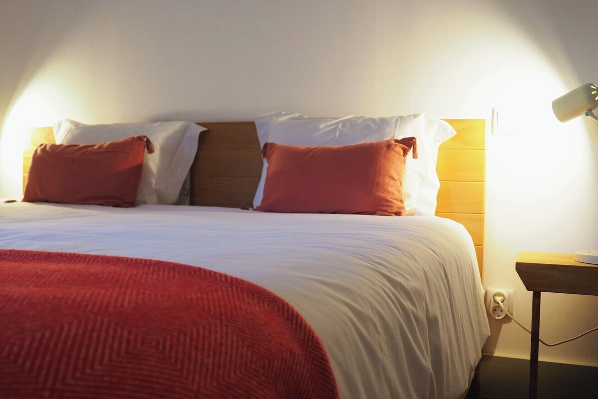 Ein ordentlich gemachtes Bett mit roter Decke und Kissen, ein Nachttisch mit Lampe und eine Steckdose.