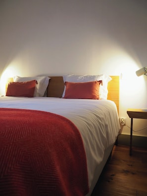 Ein ordentlich gemachtes Bett mit roter Decke und Kissen, ein Nachttisch mit Lampe und eine Steckdose.