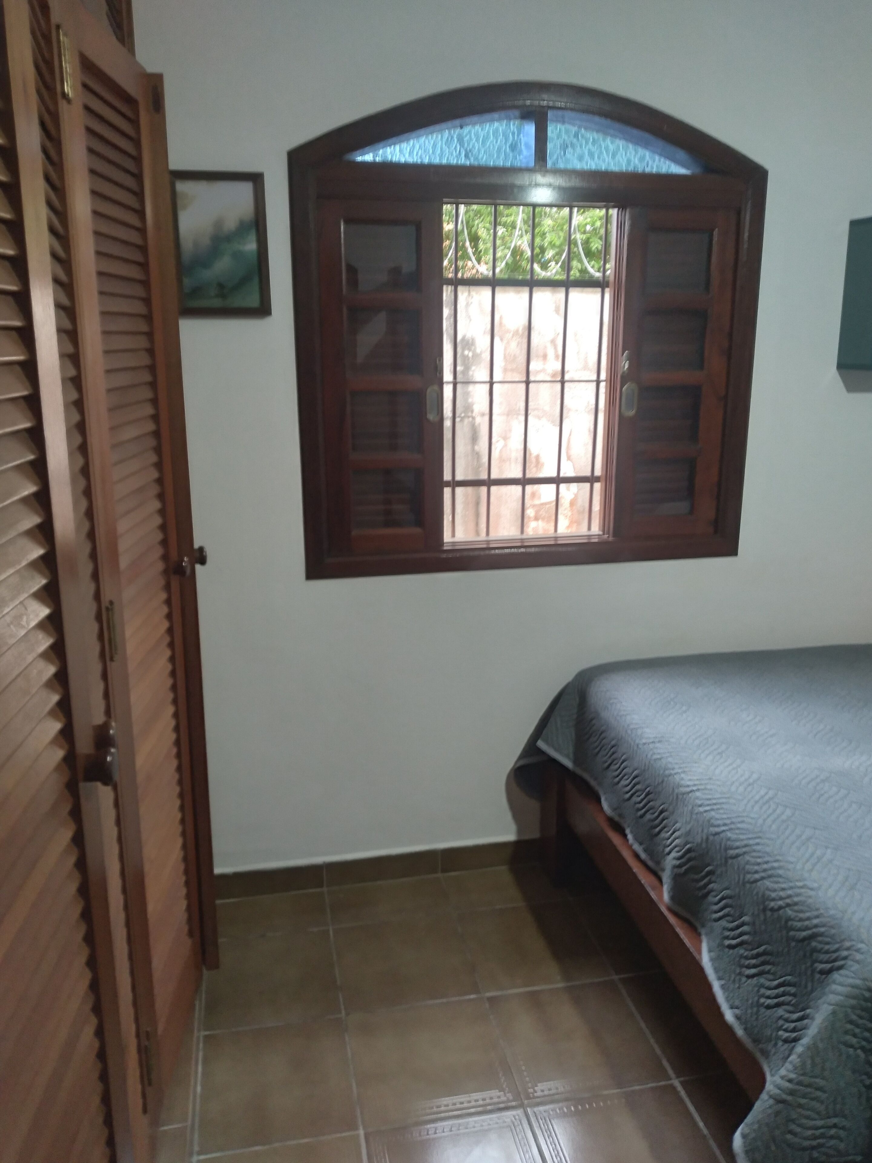 Una habitación con una cama, un ventanal con contraventanas de madera, una puerta y un suelo de baldosas.