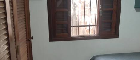 Una habitación con una cama, un ventanal con contraventanas de madera, una puerta y un suelo de baldosas.