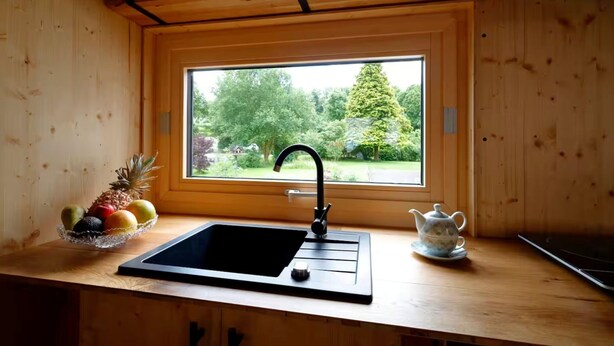 A kitchen with a wooden interior, a black sink, a window with a view of greenery, and a fruit bowl on the counter. - Tiny House Prum - Natururlaub in der Eifel (Pruem)