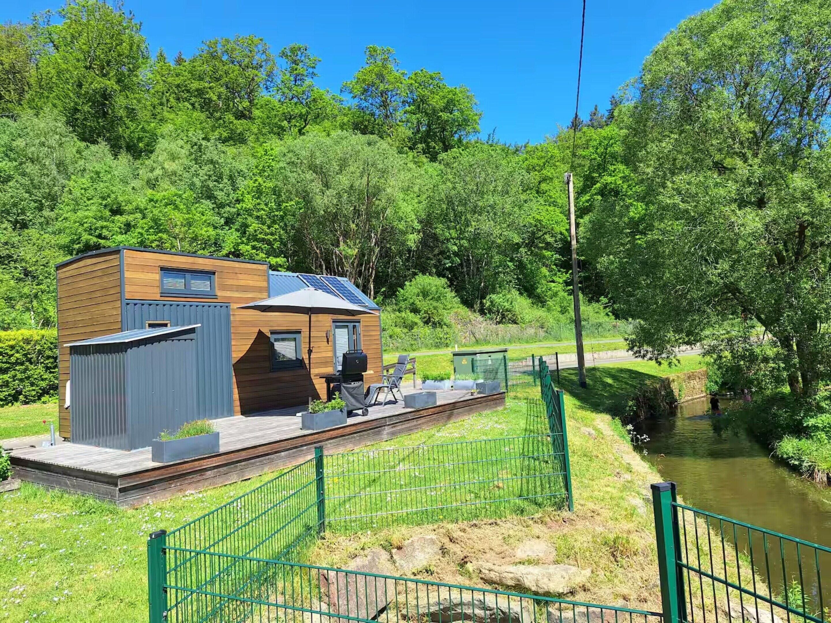Eine moderne Holzhütte mit einer Terrasse, Solaranlagen und Blick auf einen Fluss.