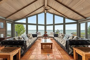 A spacious room with a high ceiling, large windows, and a view of the water. There are two leather sofas with patterned cushions and a wooden coffee table.