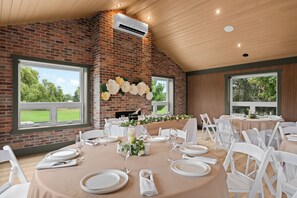 A dining area with brick walls, large windows, and a fireplace. Tables are set with white tablecloths and plates.