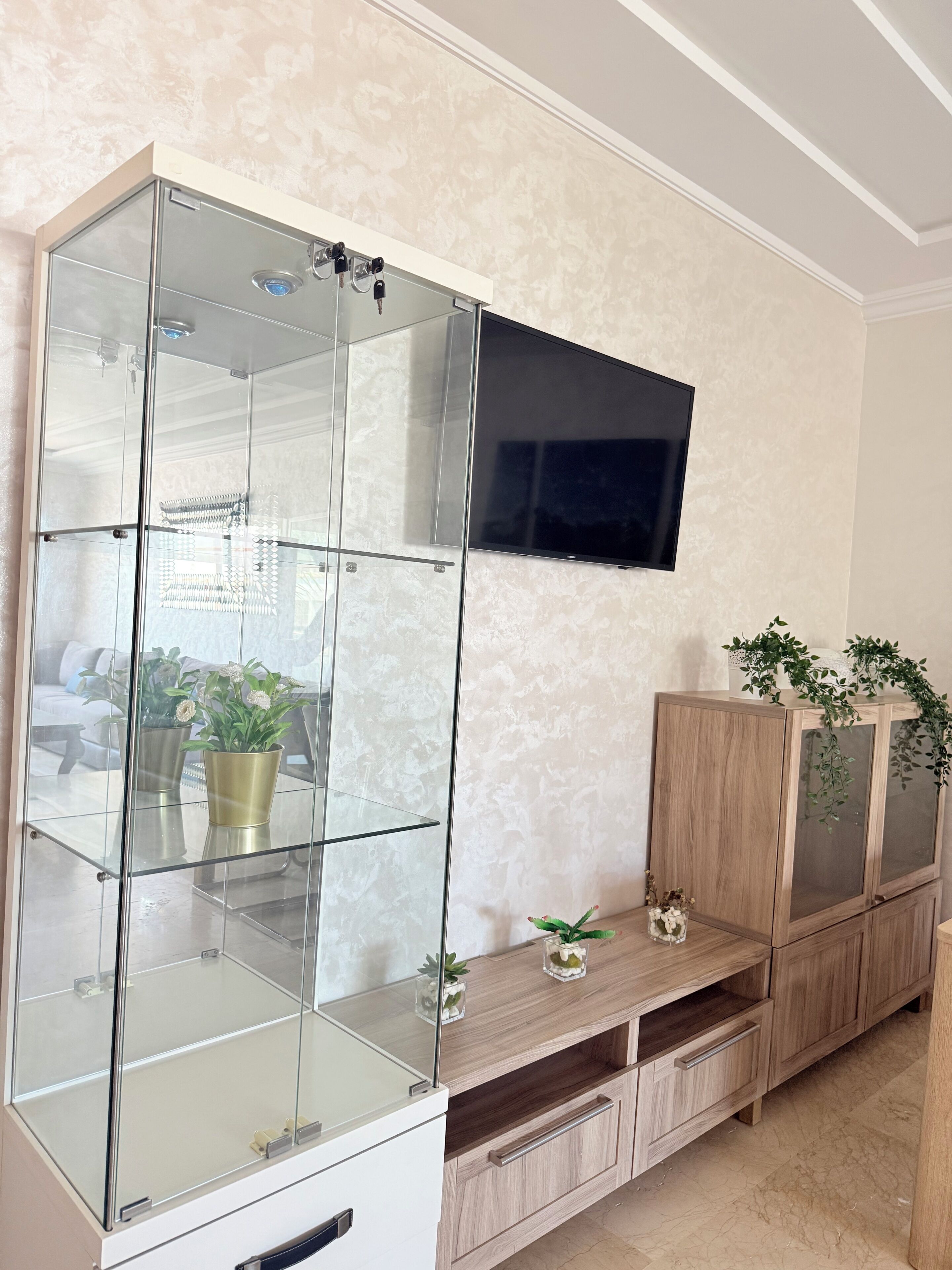 A living room with a glass-fronted cabinet, a mounted TV, and wooden furniture.