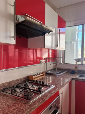 A kitchen with a gas stove, stainless steel range hood, red and white cabinets, and a window providing natural light. - Bright apartment with a pool, 5-minute walk to the beach (Témara)