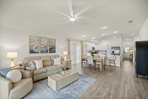 A modern living room with a sofa, coffee table, and artwork. There is a dining area with a table and chairs. The kitchen is visible in the background with cabinets and appliances. - Dog-Friendly Coastal Cottage | Golf Cart (Sumterville)