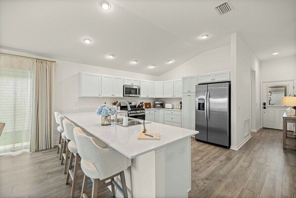 A modern kitchen with white cabinetry, stainless steel appliances, and a central island with bar stools. - Dog-Friendly Coastal Cottage | Golf Cart (Sumterville)
