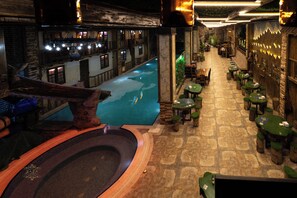 An indoor swimming pool with a stone floor, surrounded by wooden tables and chairs, and a view of a building with windows. - BazaHotel&Spa (Mil)