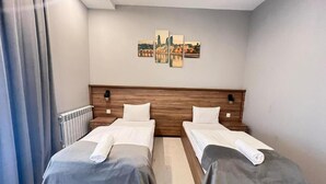 A hotel room with two beds, a wooden headboard, a cityscape artwork on the wall, and a radiator. - BazaHotel&Spa (Mil)