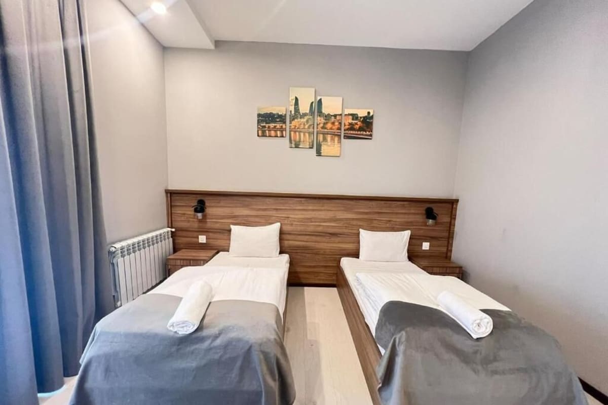 Hotellihuone, jossa on kaksi sänkyä, puinen pääty, seinällä kaupunkimaisemaa esittävä taideteos ja patteri.