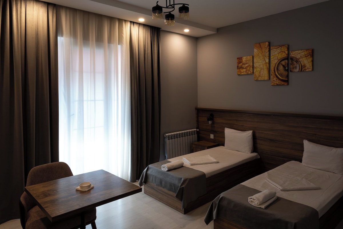 Hotellihuone, jossa on kaksi sänkyä, pöytä ja tuoli. Huoneessa on ikkuna, jossa on verhot, kattovalaisin ja seinällä taideteos.