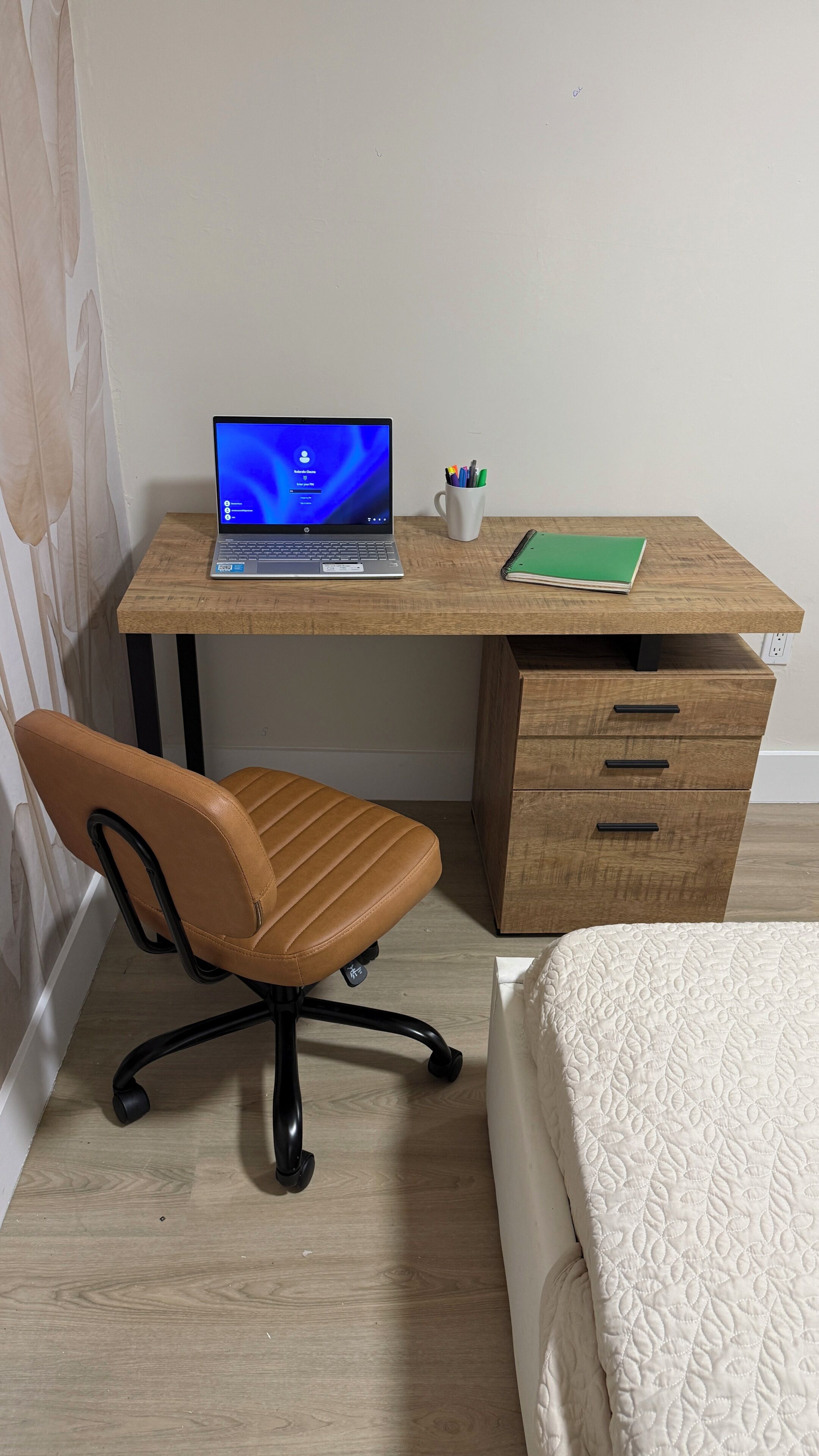 Ein Zimmer mit einem Holztisch, einem Laptop, einem Stuhl, einem grünen Notizbuch und einem Bett mit weißer Bettdecke.