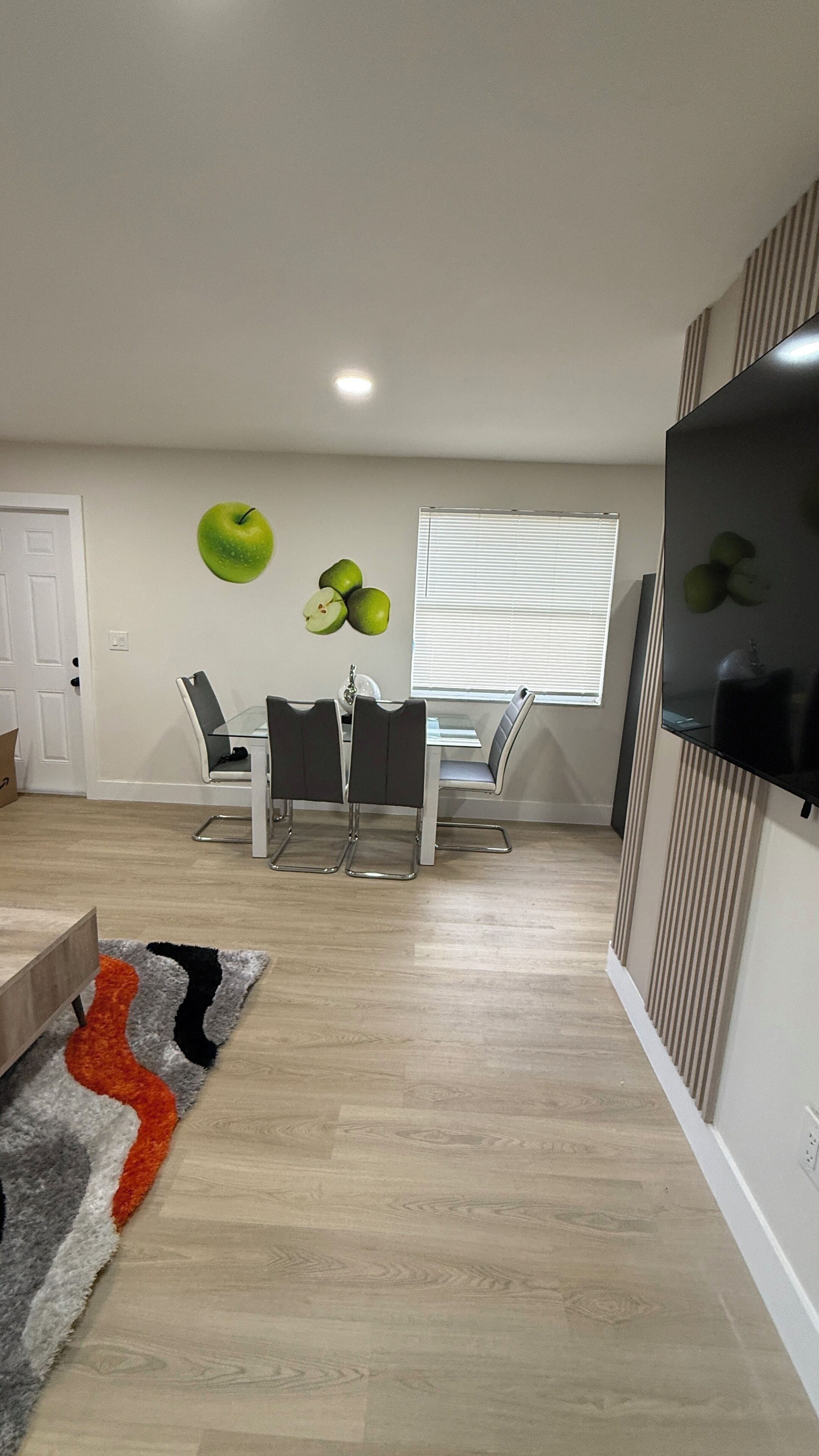 A modern living room with a dining area, a flat-screen TV mounted on the wall, and a window with blinds.