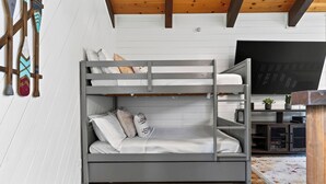 Una cama literera con escritorio y televisor en una habitación con paredes blancas y vigas de madera.