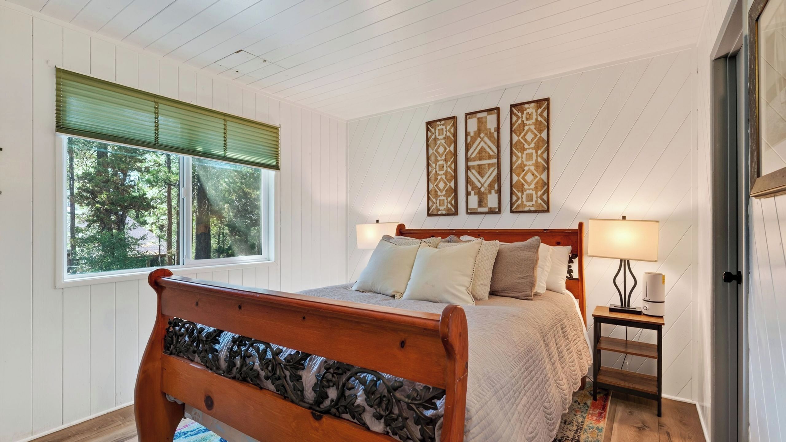Un dormitorio con cama de madera, una ventana con persianas, una mesita de noche con lámpara y un cuadro con motivos decorativos en la pared.
