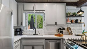 A modern kitchen with white cabinetry, a built-in oven, a coffee maker, and a window with a view of greenery. - Balboa Big Bear by Avantstay Hot Tub + Game Room + Firepit + Putting Green Near Bear Mountain (Big Bear Lake)