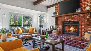 A cozy living room with a fireplace, comfortable seating, and a view of the outdoors. - Balboa Big Bear by Avantstay Hot Tub + Game Room + Firepit + Putting Green Near Bear Mountain (Big Bear Lake)
