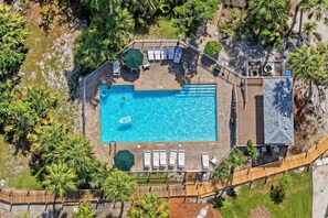 An aerial view of a rectangular swimming pool surrounded by lounge chairs and umbrellas, with a wooden deck and a covered area with air conditioning units. - 1 Block to Beach | Garage Game Room | Community Pool | Beach Gear | Pet Friendly (Holmes Beach)