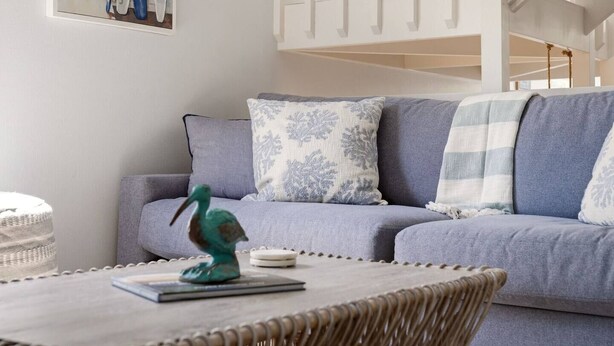 Ein modernes Wohnzimmer mit einem grauen Sofa, gemusterten Kissen, einem kleinen Tisch mit einer Dekoration und einem Korbstuhl.