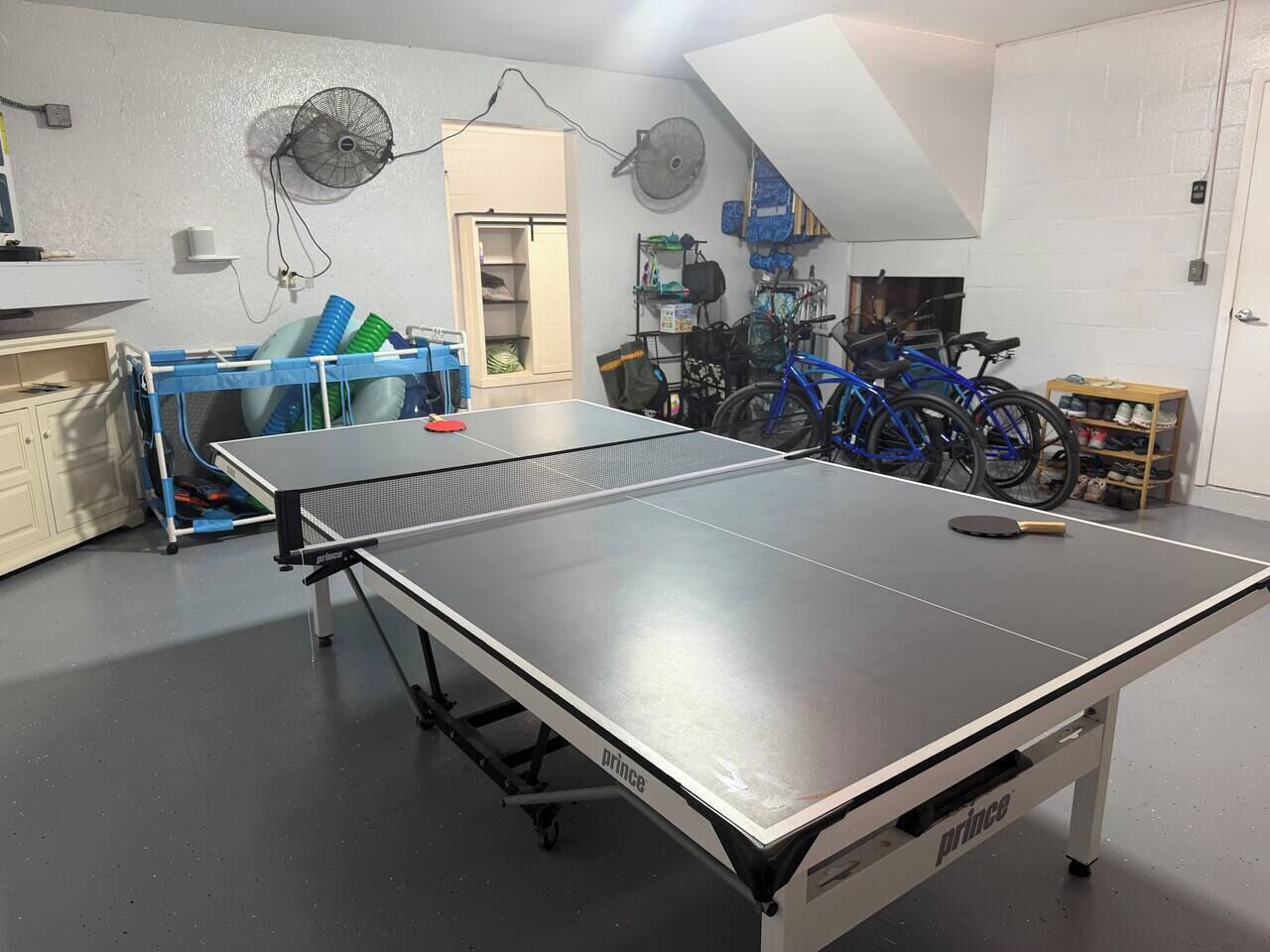 Une table de ping-pong dans une pièce où se trouvent des vélos et un ventilateur.