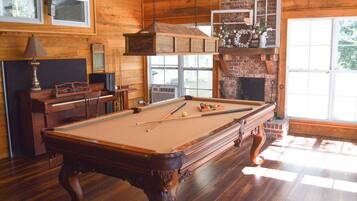 A pool table with billiard balls, a piano, and a fireplace in a wooden room with large windows.