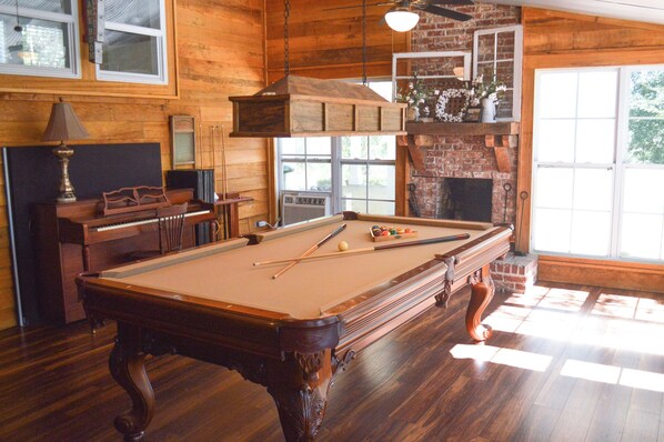 A pool table with billiard balls, a piano, and a fireplace in a wooden room with large windows.