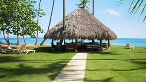 A thatched-roof hut with a concrete pathway leading to it, surrounded by palm trees and beach chairs on a grassy area. - Villa Pura Vida 4BR private Villa with large Pool & Bungalow (Las Terrenas)