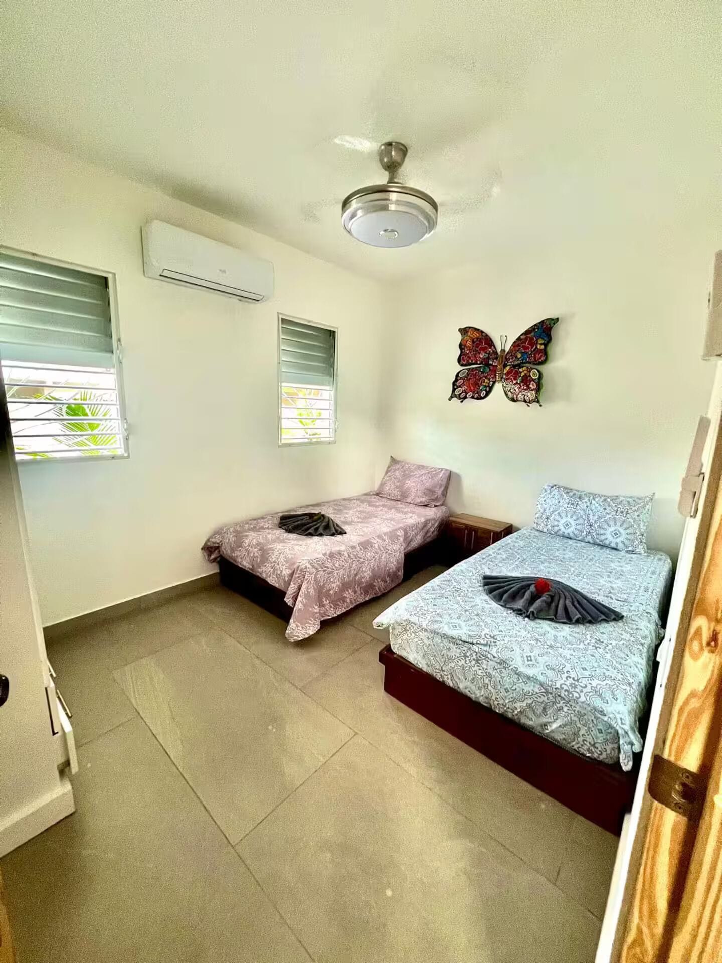 Um pequeno alojamento de um quarto, com duas camas, ventoinha no teto e uma decoração com borboletas.