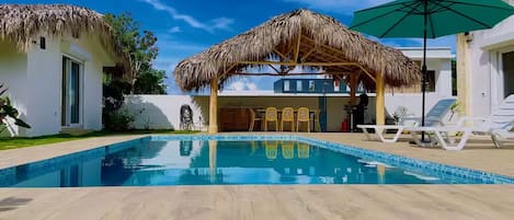 Une piscine dotée d’une structure à toit de chaume, de chaises longues et surplombée d’un ciel bleu azur.
