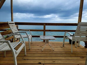 A wooden deck with white metal chairs and a small table, overlooking a body of water. - JV BEACHFRONT APARTMENT: YOUR DREAM OCEANVIEW SPOT (Añasco)