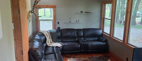 A log cabin interior with a black leather sectional sofa, a ceiling fan, and a large window offering a view of the outdoors.