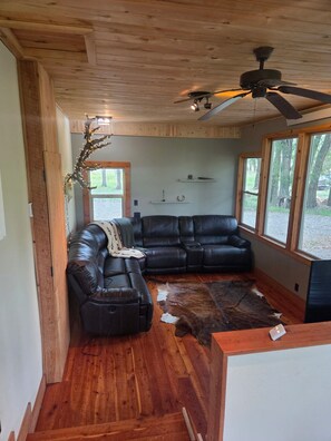A log cabin interior with a black leather sectional sofa, a ceiling fan, and a large window offering a view of the outdoors. - House just minutes from the Yellowstone river. (Livingston)