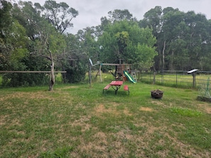 A playground with a slide, swings, and a picnic table in a grassy area with trees in the background. - House just minutes from the Yellowstone river. (Livingston)