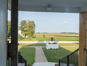 A view from a porch with a pathway leading to a bell in a white structure, surrounded by green fields and a large tree.