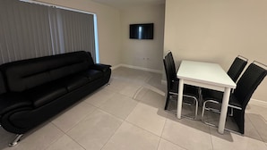 A living room with a black sofa, a white dining table with chairs, a flat-screen TV mounted on the wall, and a ceiling light fixture. - High Wycombe Apartments (High Wycombe)