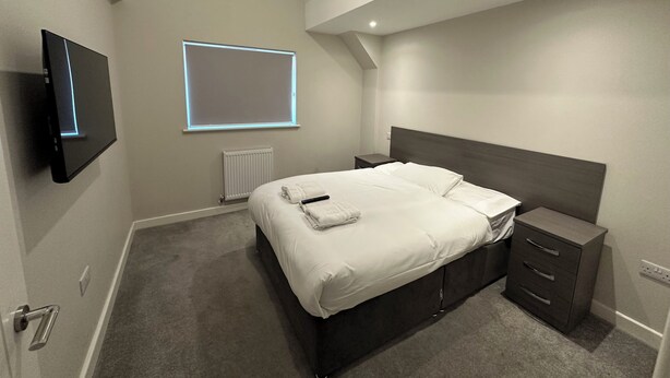 A bedroom with a bed, a bedside table, a nightstand, a TV mounted on the wall, and a window with blinds. - High Wycombe Apartments (High Wycombe)