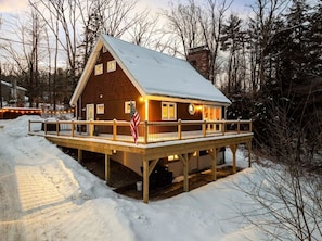 A wooden cabin with a deck, surrounded by snow and trees.