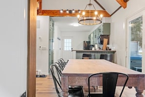 A dining area with a long wooden table and chairs, a chandelier, and a view of the kitchen and outdoor area.
