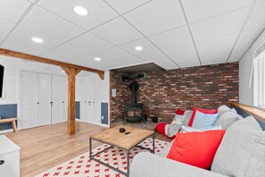 A cozy living room with a brick wall, a sofa with red and grey cushions, a wooden coffee table, and a white ceiling with recessed lighting.